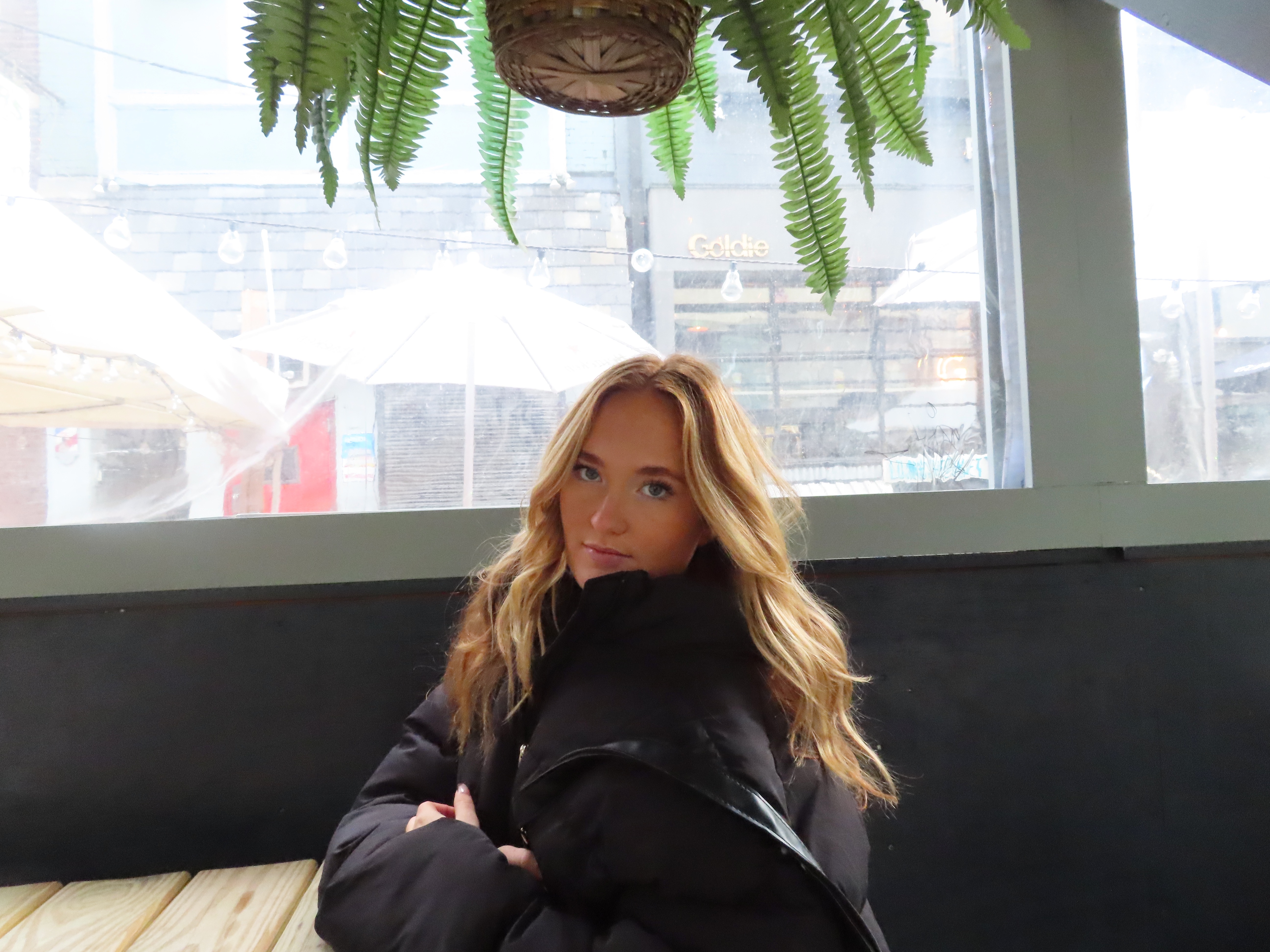 This is a rectangle image of Grace sitting at a wooden table in the city. She is wearing a black puffer jacket with a black leather purse around her shoulder. Her hair is red with blonde highlights, and it is down and slightly curled. She has mascara and light face makeup on. There is a green hanging plant above her in a brown basket, and the back ground is washed out. The back ground has a white umbrella and a couple shops that are visible.