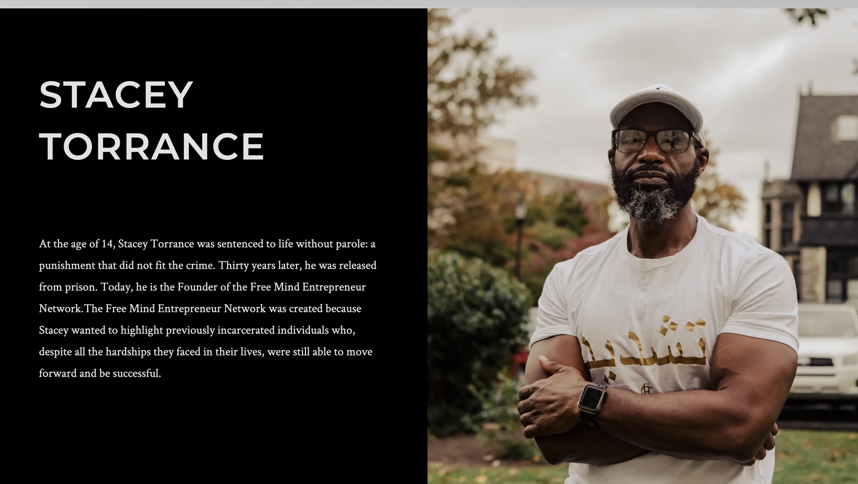 This is a screenshot photo of a wesbite. The website is called Free Mind Entrepreneur Network. The website page is a black ground with white writing. Included on the page is a photo of the founder of the organization. The founder is standing with his arms crossed, and he is wearing a white t shirt with gold writing. He is also wearing a white hat and black glasses. His beard hair is black mixed with grey. He is also wearing a black apple watch. The back ground is fall colored trees and bushes, as well as a brown building with a white car infront of it. 